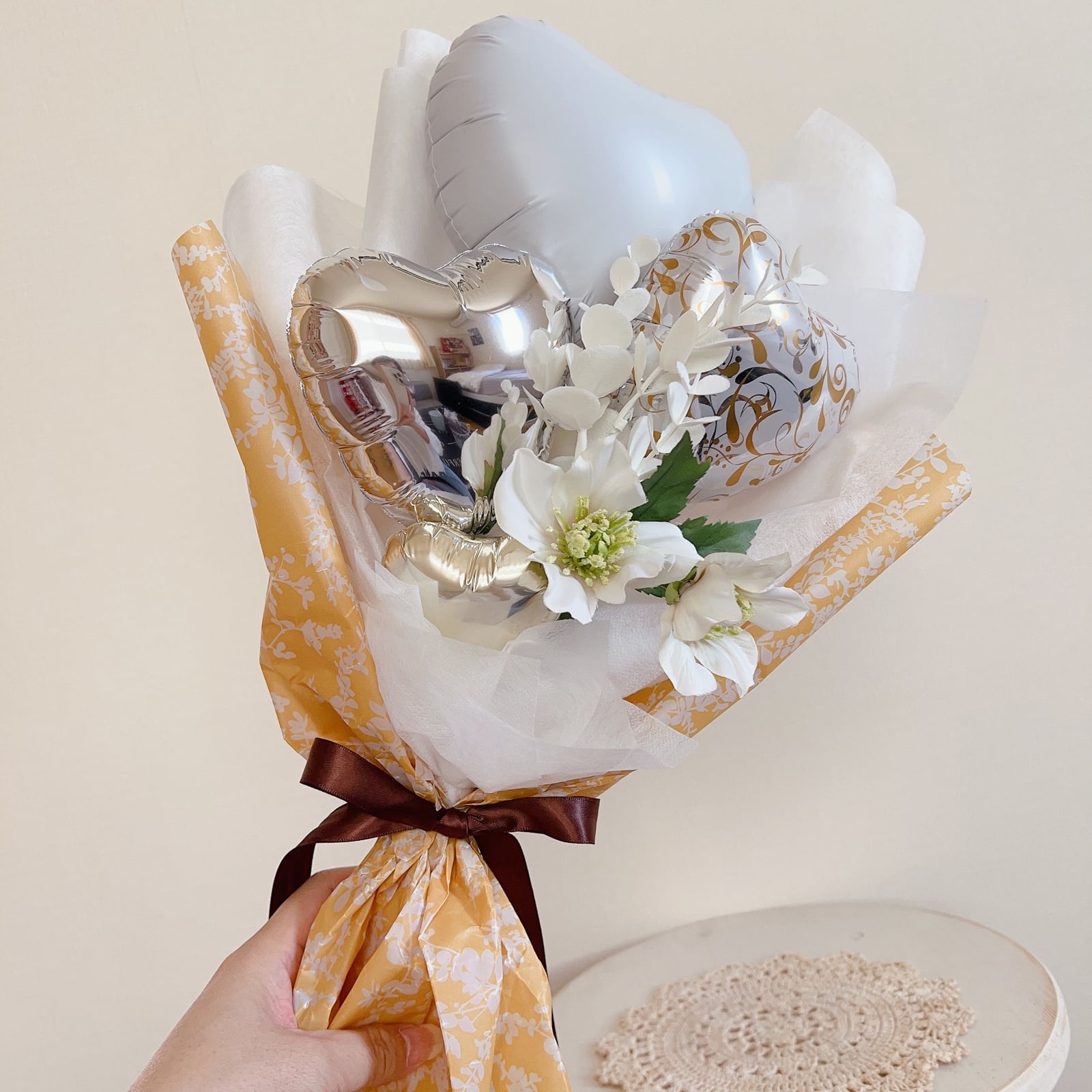白い花と白・シルバーのバルーンをゴールドの包装でまとめた小ぶりなブーケ